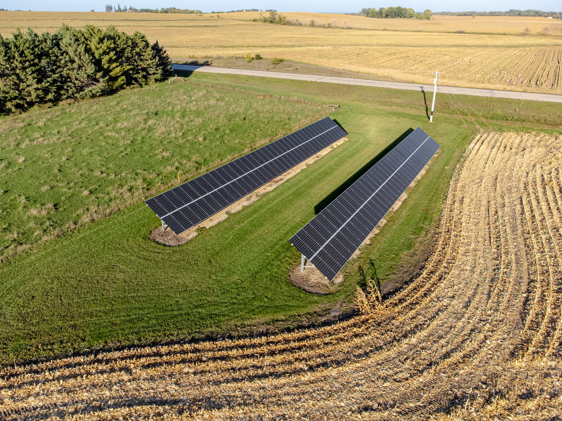 Aerial landscape photograph showing rows of solar panels beside cultivated farmland and a tree line under a partly cloudy sky.
