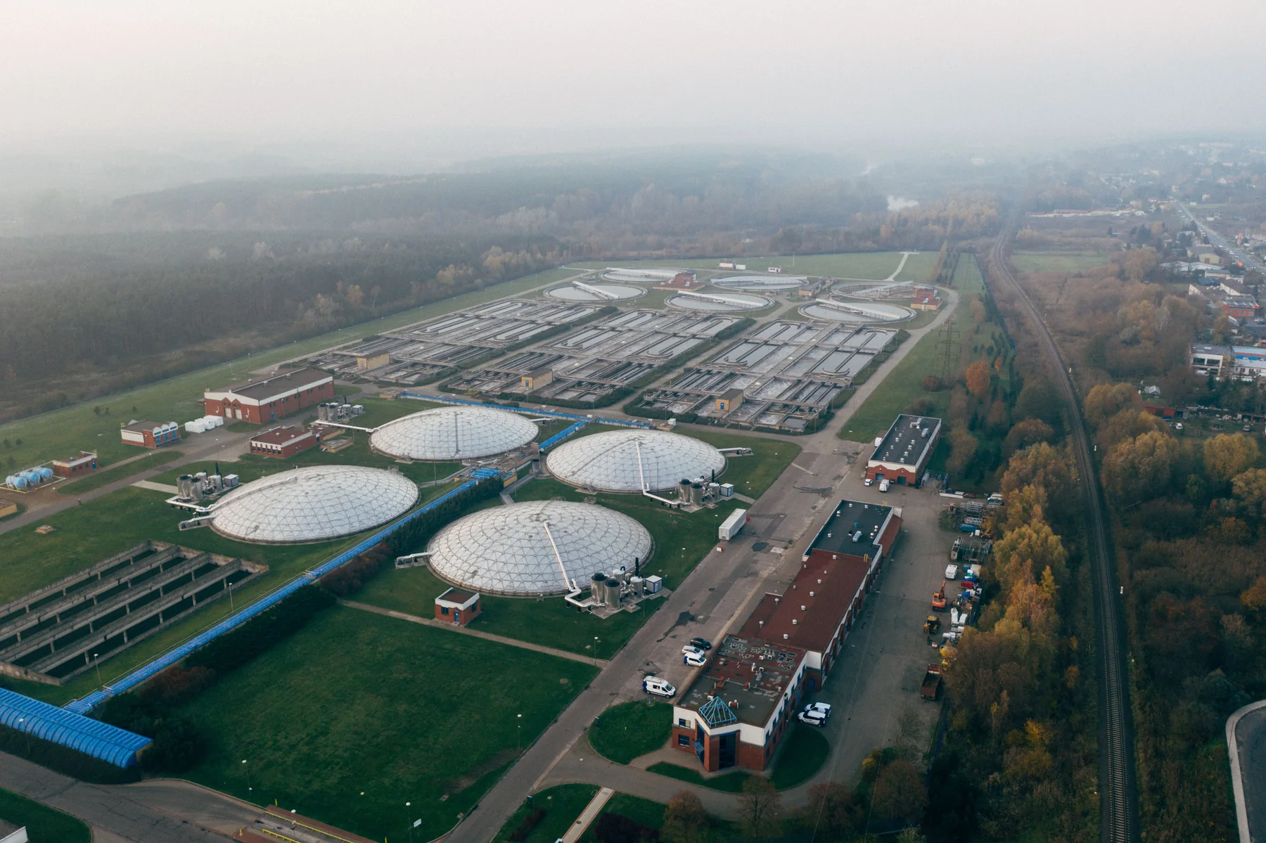 Aerial landscape photograph of a wastewater treatment facility with rectangular settling basins, circular tanks, service roads and surrounding green fields.