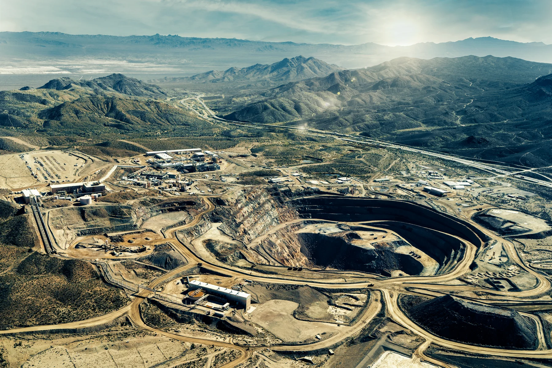 Aerial landscape photograph of a large open-pit rare-earth mining and processing complex, with terraced earthworks, roads, industrial buildings, ponds, and surrounding mountain terrain.