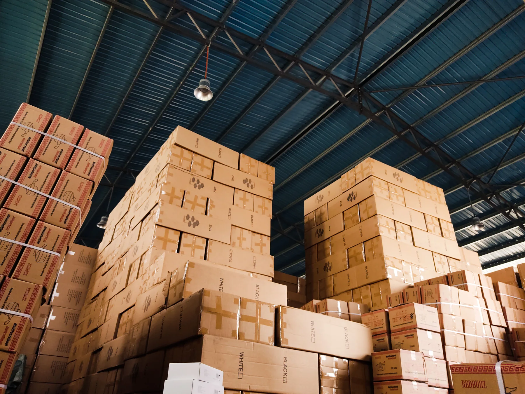 High stacks of plain cardboard boxes arranged in a warehouse beneath a blue metal ceiling, illustrating responsible sourcing and storage.