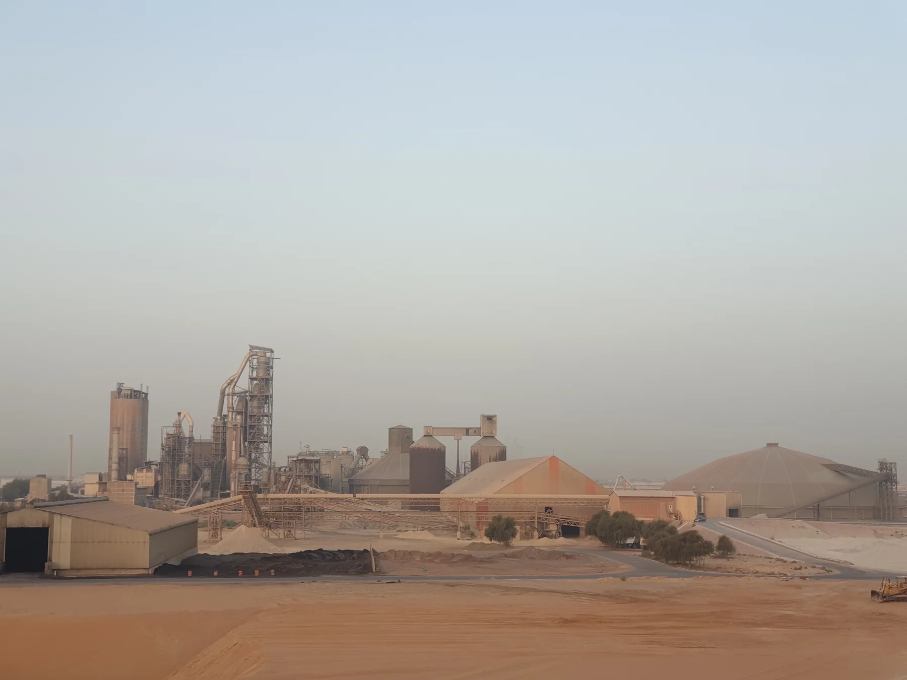 Landscape photograph of a large industrial complex with processing towers, storage buildings, covered stockpiles, access roads and sandy ground under a hazy sky.