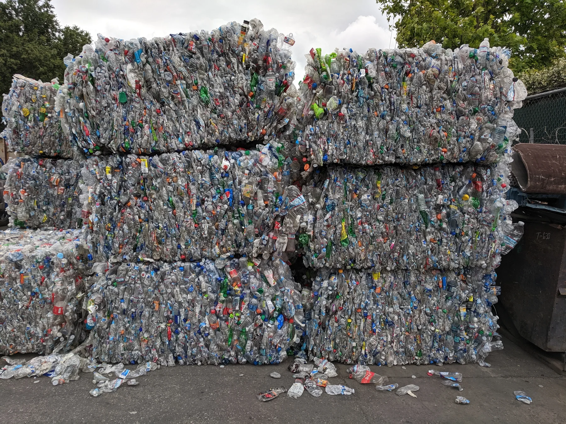Large bales of crushed plastic PET drink bottles stacked at a recycling facility, viewed outdoors.