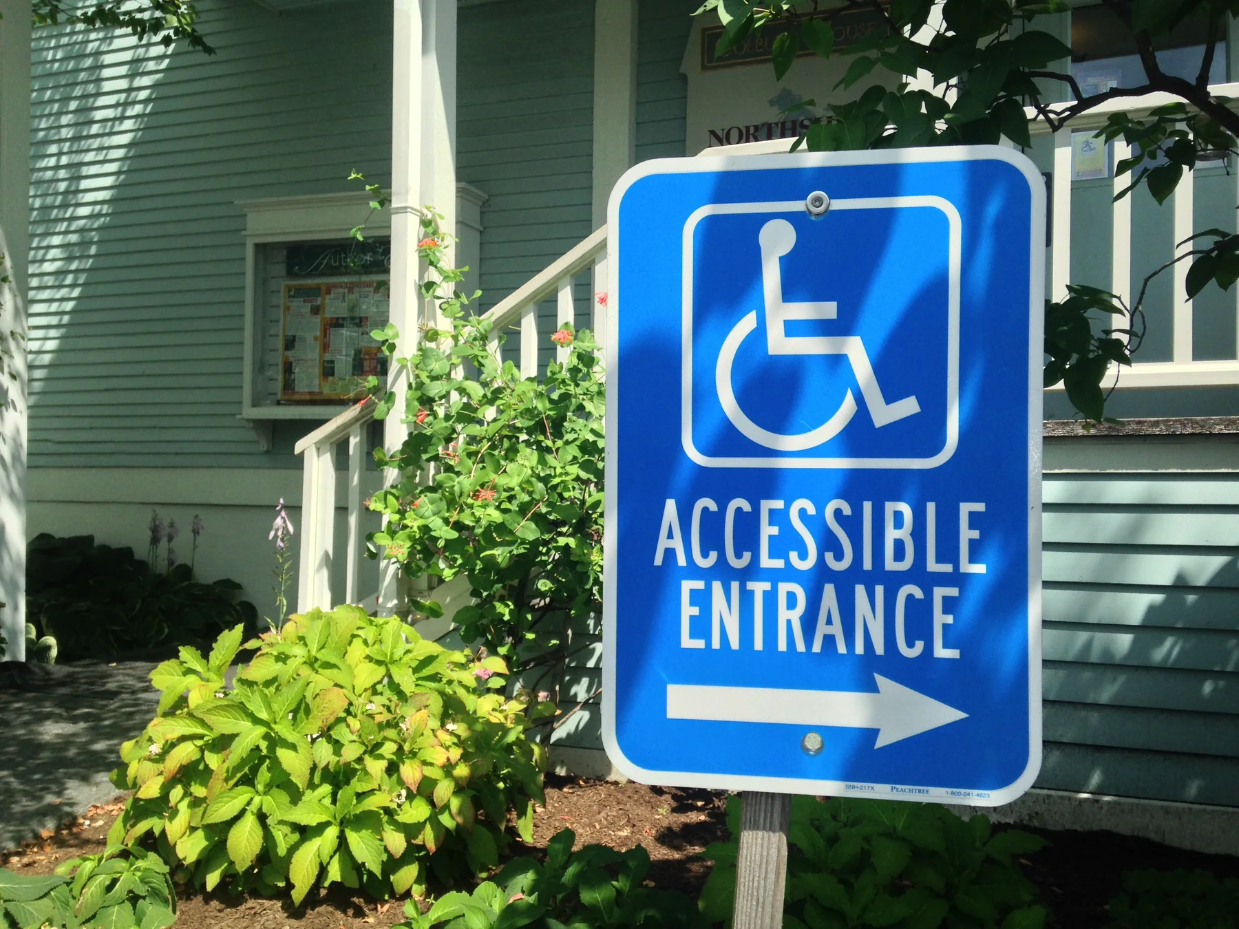 Landscape photograph of a blue accessible-entrance sign beside a landscaped building approach, illustrating a physical-access consideration for inclusion training.