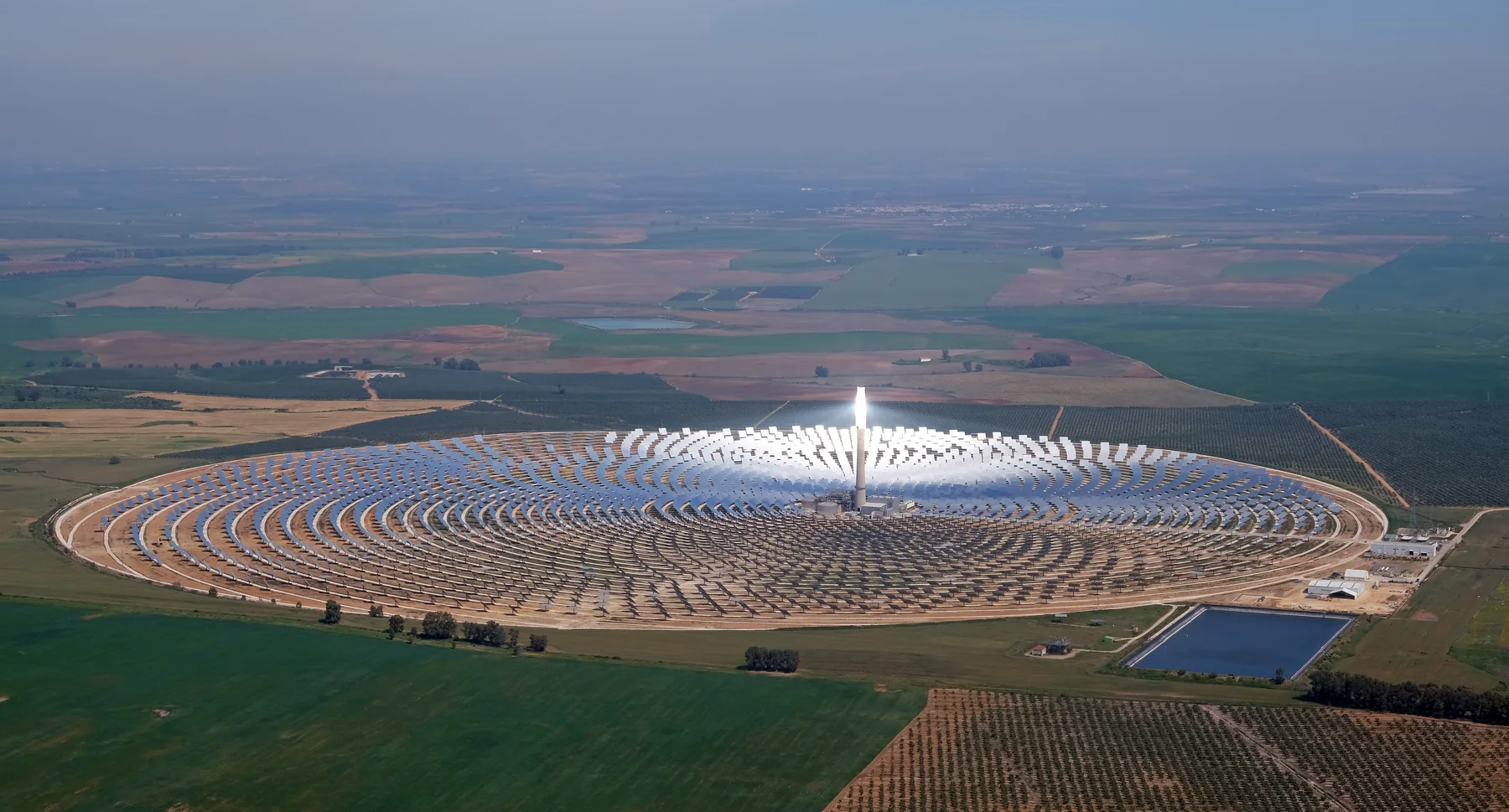 Aerial landscape view of the Gemasolar thermosolar power plant, with a central solar-tower installation and surrounding industrial solar field under a partly cloudy sky.