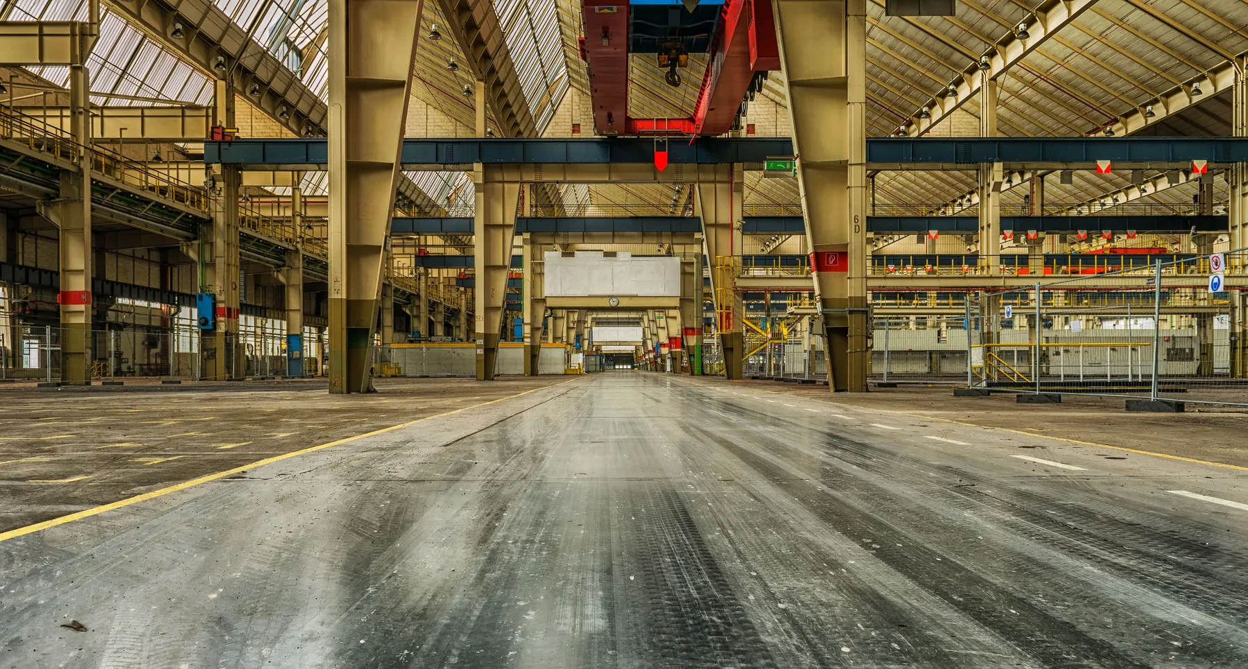 Wide view of an empty industrial warehouse interior with steel roof trusses, overhead cranes, concrete floor, and metal structures.