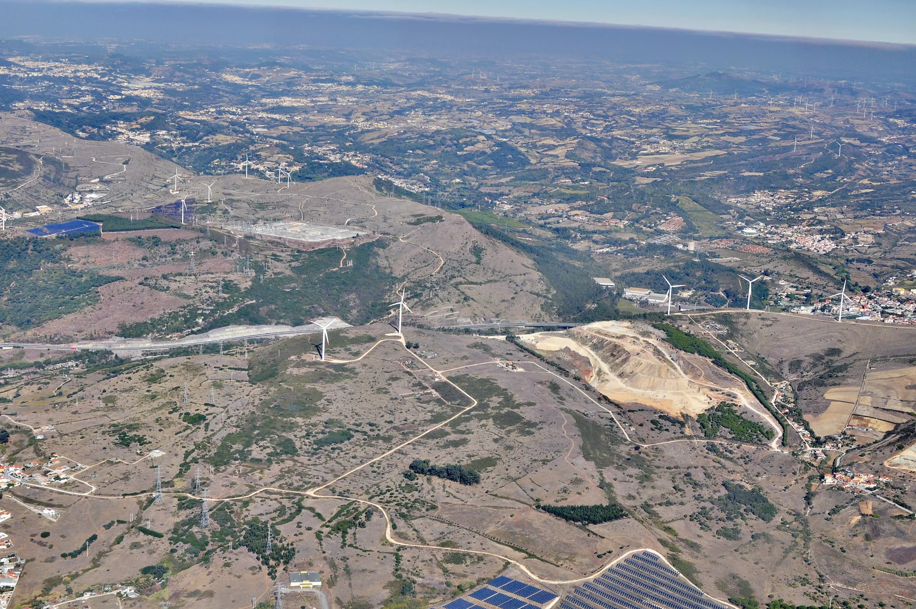 Aerial landscape showing rows of solar panels and wind turbines across agricultural fields under a blue sky.