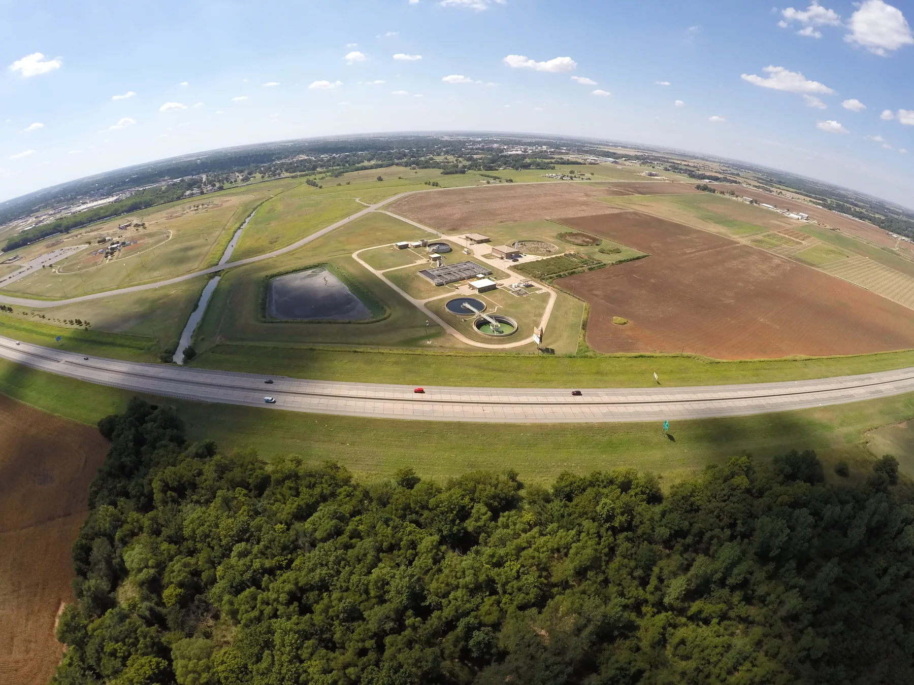 Aerial view of circular wastewater treatment ponds, rectangular treatment beds, service buildings, roads, and adjacent agricultural fields.