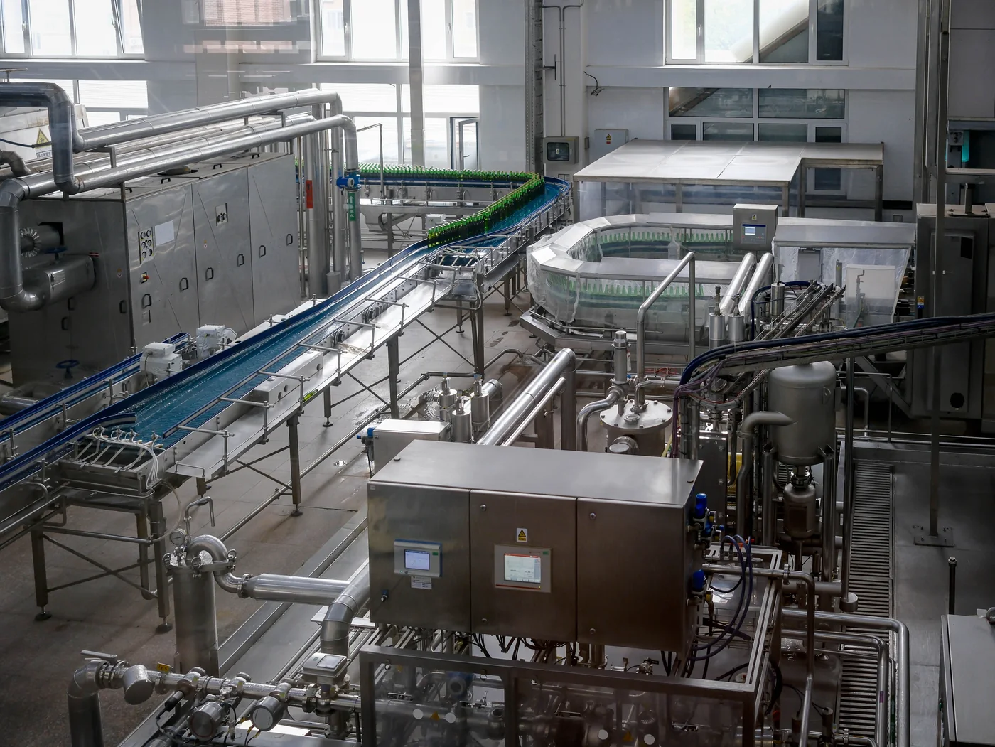 Bright automated beverage bottling factory interior with green bottles on curved conveyor belts amid stainless-steel machinery and pipes.